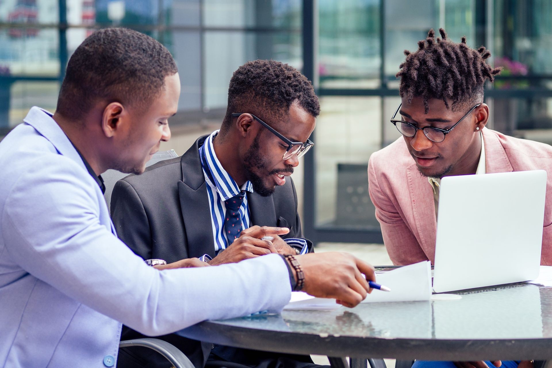 A group of three stylish African businessman partners entrepreneurs in fashion business suits meeting sitting at table sign securities with laptop in a summer cafe outdoors